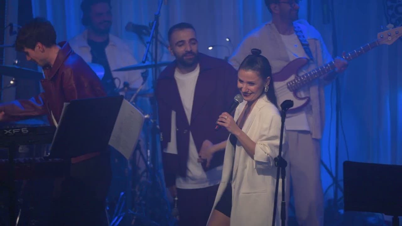 A singer in a white blazer smiles as she holds a microphone on a blue-lit stage, with a keyboard player, a drummer, a man in a purple jacket and a bass player behind her, in Ankara Echoes' live video of 'Beni Al'