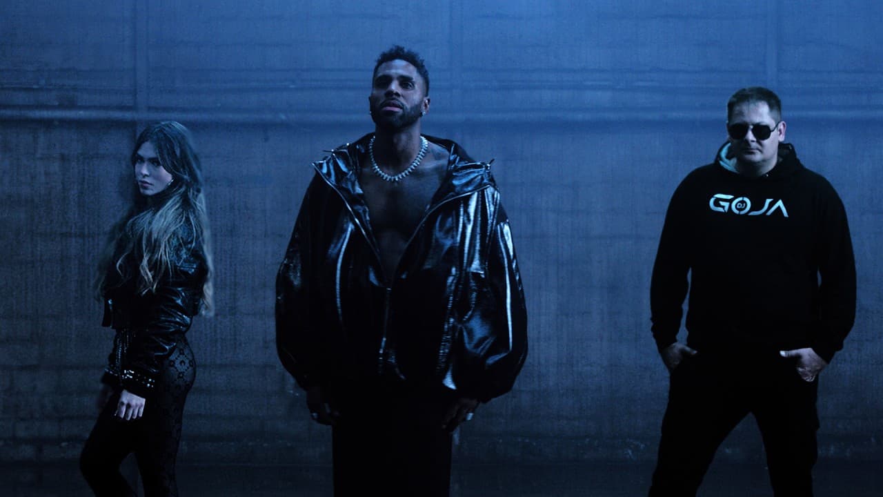 DJ Goja in a black GOJA hoodie and sunglasses stands beside Jason Derulo in a shiny black jacket and a woman in black against a blue concrete wall, in the 'Mi Chico' video