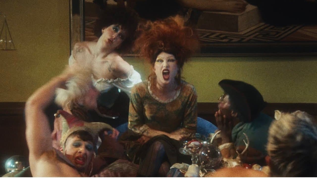 Haute, with a towering mass of red curls, white face paint and red lips, shouts from a sofa among costumed dancers in a dim, crowded room in the 'Shy Girl' music video, while a man in glasses and a dark cap raises his hands to his face beside her