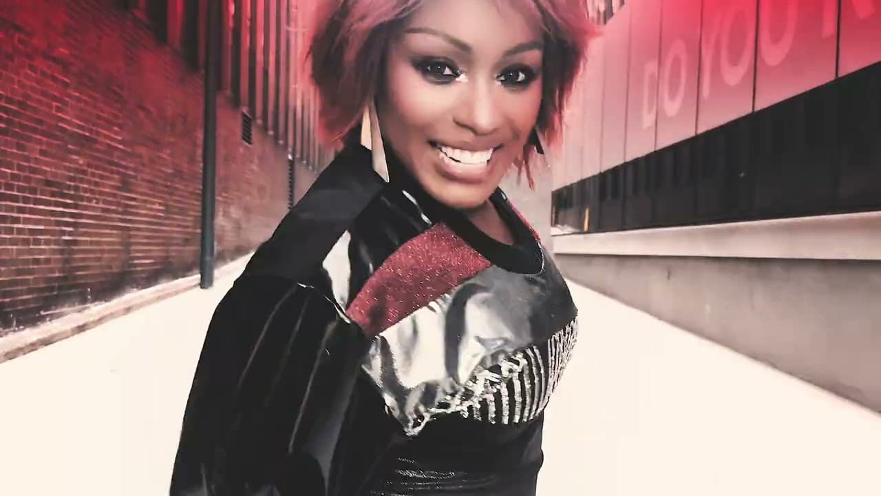 Ultra Naté 'Movin' To The Sun' MV capture: the singer smiling in a red-lit alley between brick and glass walls
