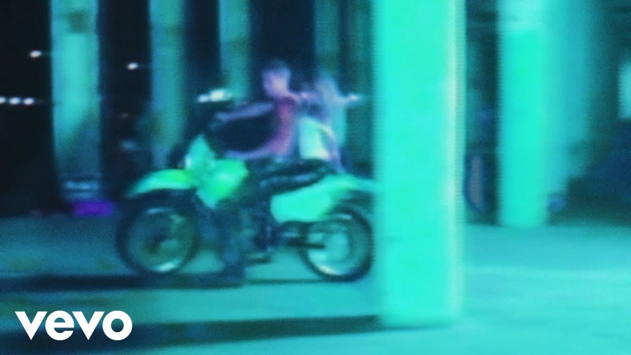 Two people ride a green dirt bike at night through a colonnade lit teal and violet, from the official “BbY WOW” visualizer