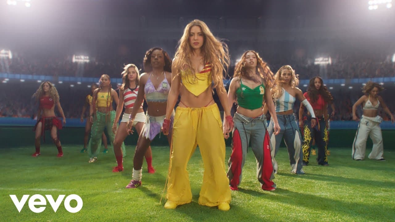 Shakira & Burna Boy 'Dai Dai' MV capture: Shakira in yellow trousers leads a line of dancers in bright football-shirt colours across a stadium pitch