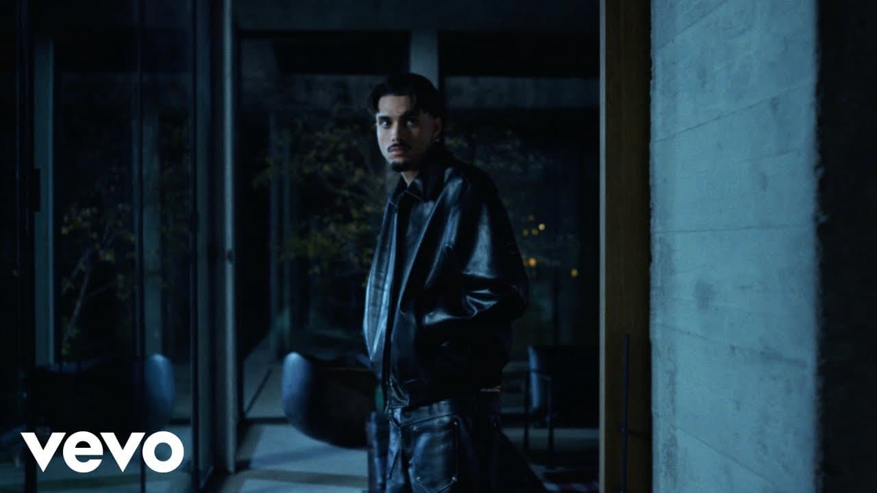 RnBoi in a black leather jacket looks back over his shoulder inside a concrete-and-glass house at night in the 'J'assume' clip