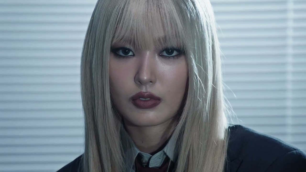 KATSEYE 'Animal' MV capture: a member with long platinum hair, heavy eye make-up and dark lipstick looks straight at the camera in a suit jacket, a window blind behind her