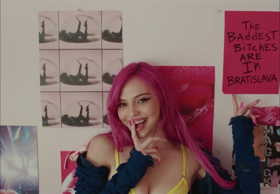 ADÉLA in a yellow top and a navy cardigan, a finger to her lips, in front of a bedroom wall of posters and a hand-lettered pink sign naming Bratislava, in the music video for 'Ain't In LA'