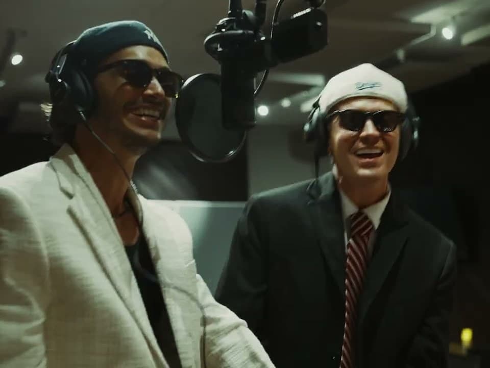 Jamie Miller and Antonio Cipriano, both in dark sunglasses, laugh as they share a studio microphone in the 'Babydoll' duet video; one wears a cream blazer and navy baseball cap, the other a black suit, striped tie and white cap