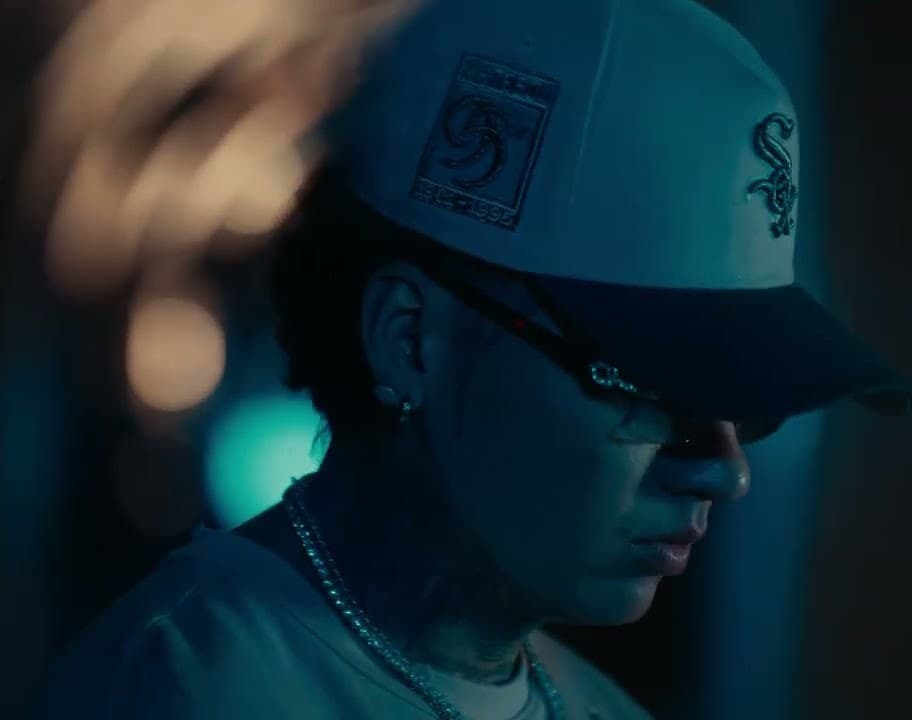 BLESSD x ANUEL AA 'CAPCANA COCOA' MV capture: a close-up of a rapper in a white cap and sunglasses, head turned down, with blurred amber lights behind him