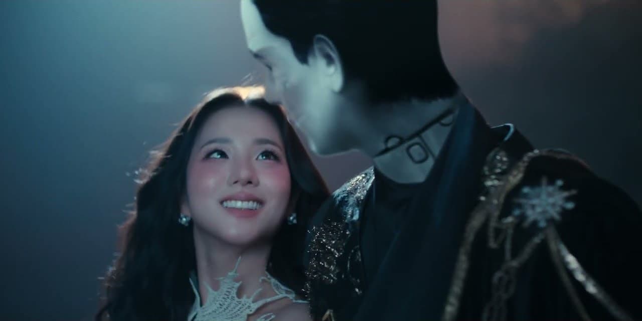 JISOO 'CLICK' MV capture: JISOO smiles up at a pale figure in a dark, gold-embroidered jacket who leans over her in blue light