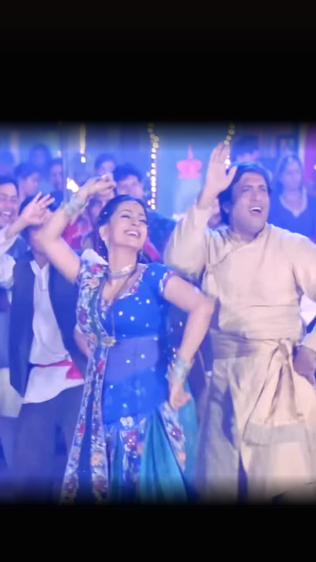 Juhi Chawla in a blue embroidered lehenga and Govinda in a cream kurta dance with their arms raised before a crowd under strings of lights in the 'Aayi Hai Diwali Suno Ji Gharwali' song sequence