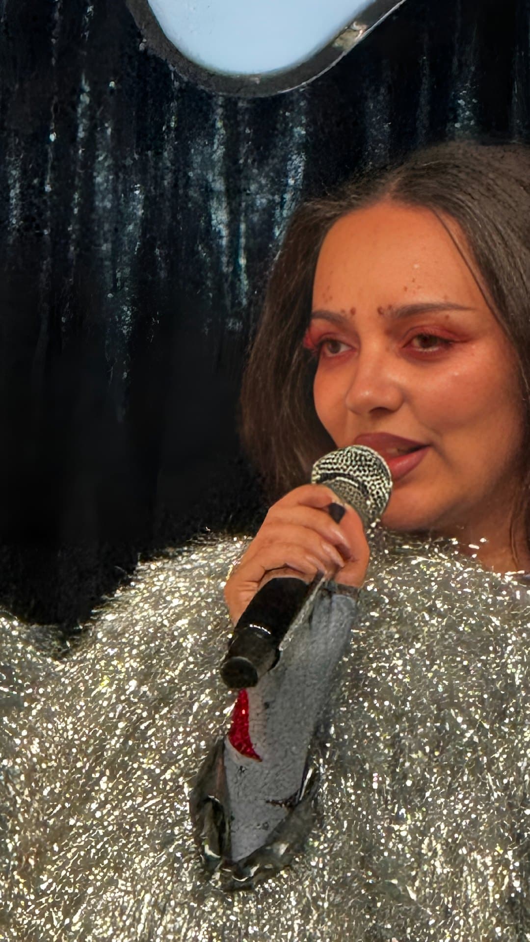 JADE, in a silver tinsel outfit with red eye make-up and painted freckles across her brow, sings into a hand-held microphone on a stage in Brussels in March 2026