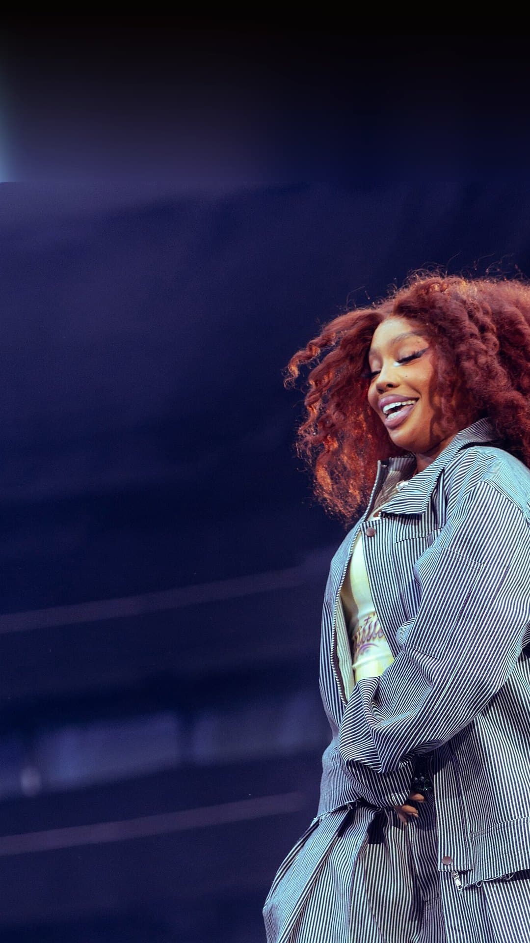 SZA, with long curly red hair and a grey striped jacket and wide trousers, smiles with her eyes lowered as she crosses the stage against a dark blue stadium in London in July 2025