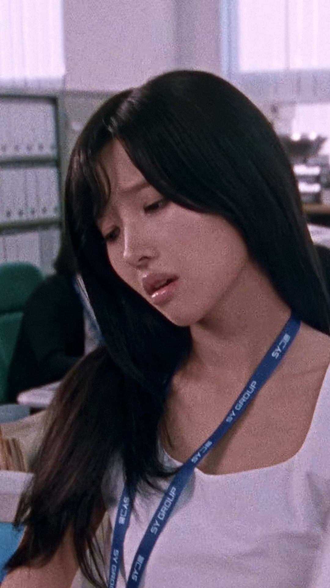 SOYEON, in a white dress with a company lanyard, looks away wearily in the office scene of the video for 'I'm gonna TOESA'