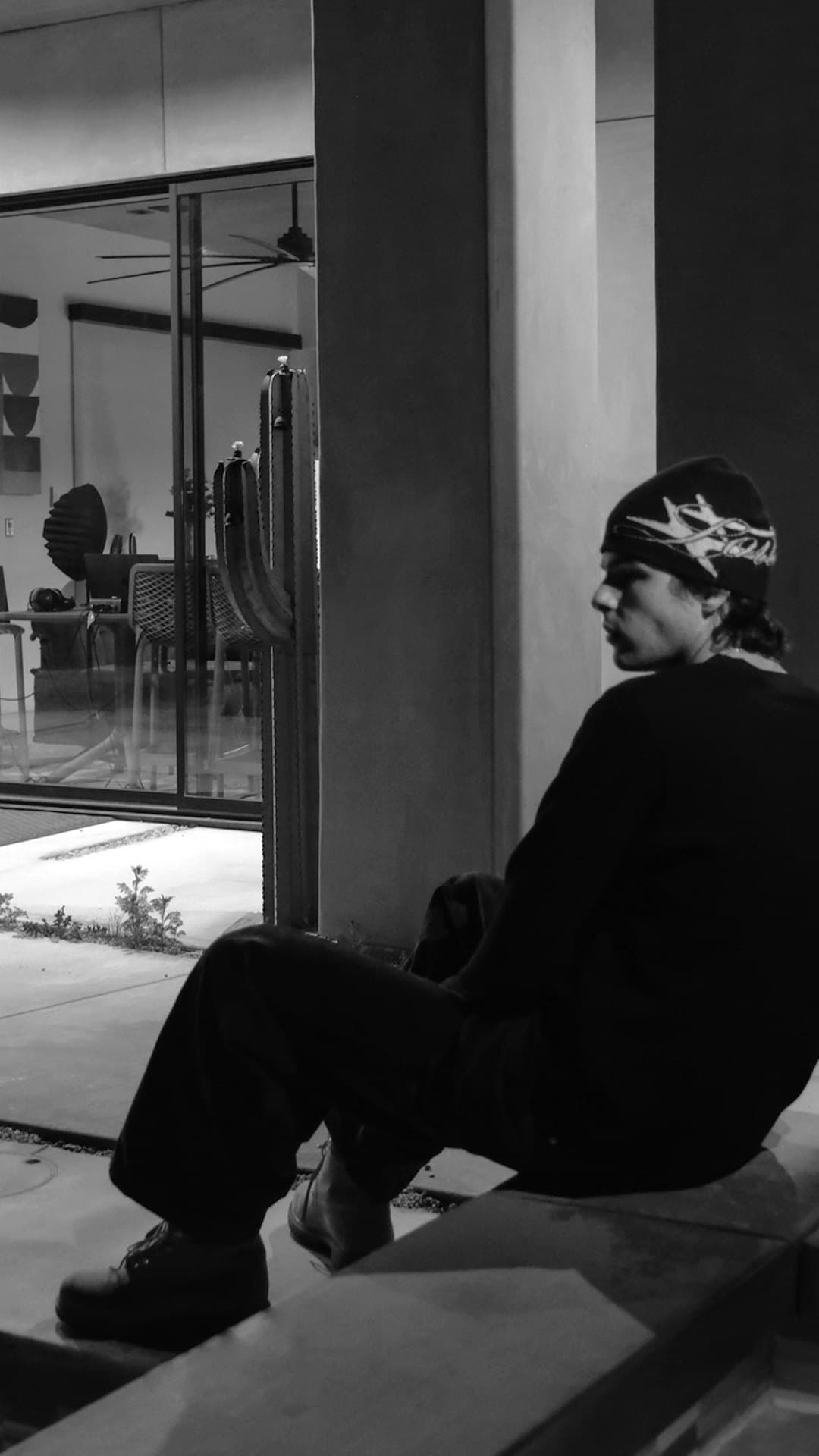 A young man in a dark beanie and dark clothes sits alone on a low ledge outside a lit glass-walled house at night, in the black-and-white photograph on the cover of Thekidszn's single 'IMPULSIVE'