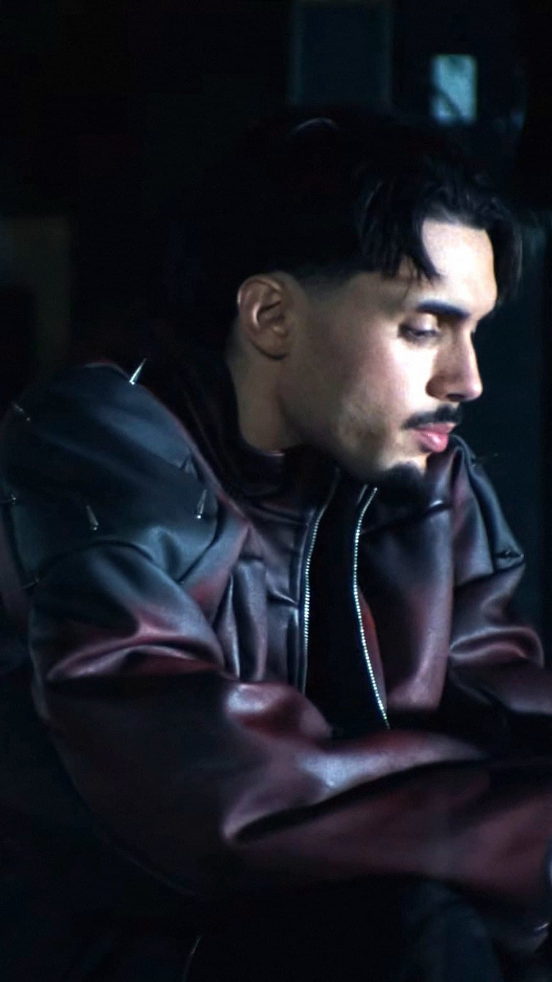 RnBoi, in a dark red leather jacket, looks down with his mouth closed in a dim room in the 'J'assume' clip