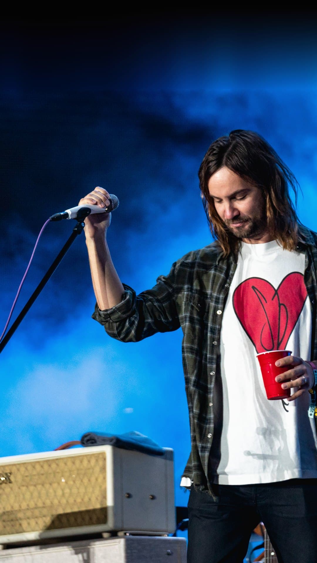 Kevin Parker of Tame Impala, in a plaid shirt over a white T-shirt with a red heart, holds up a microphone and a red cup and looks down, against blue stage smoke at Flow Festival in Helsinki in 2019