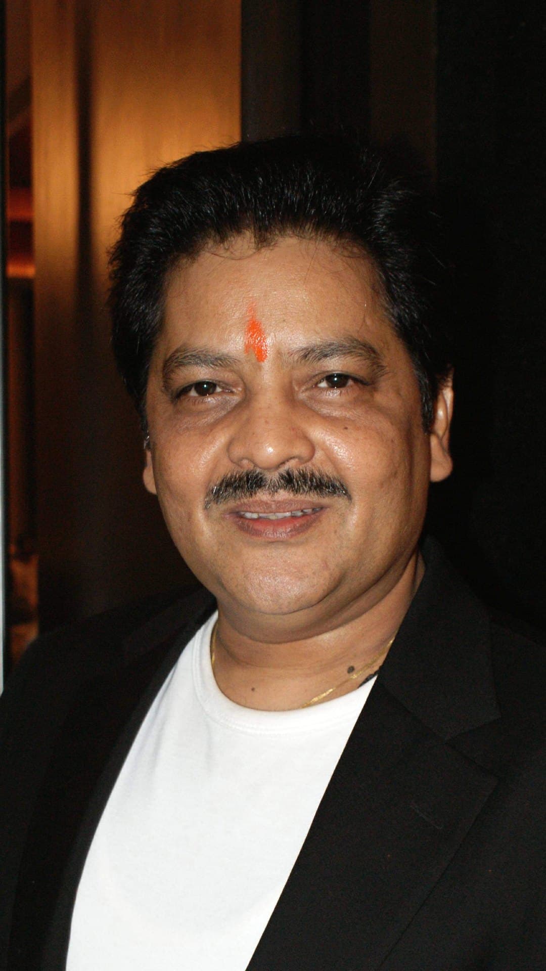 Udit Narayan, in a black blazer over a white T-shirt with a tilak on his forehead, smiles at the camera