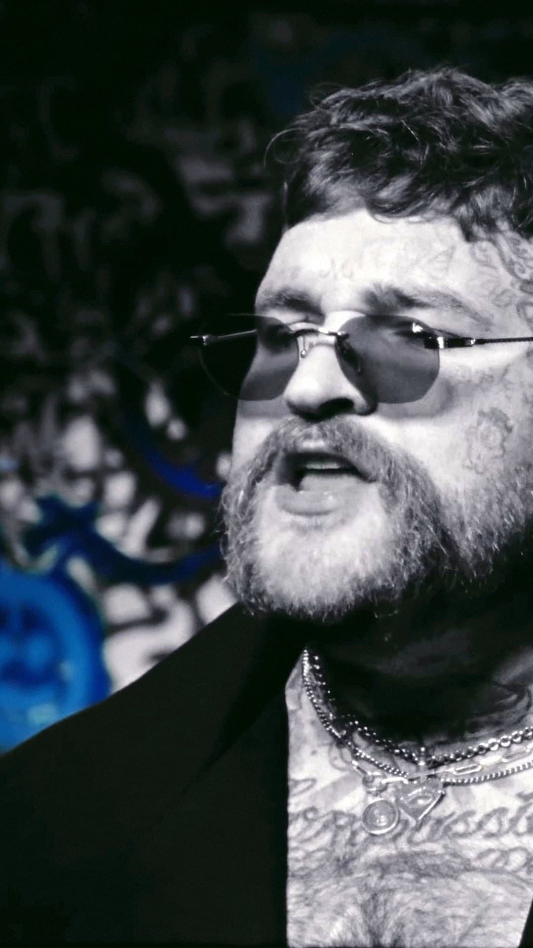 Teddy Swims, bearded, with tattoos on his face and neck, tinted rimless sunglasses and chains, sings in black and white in a close-up from the 'Mr. Know It All' video