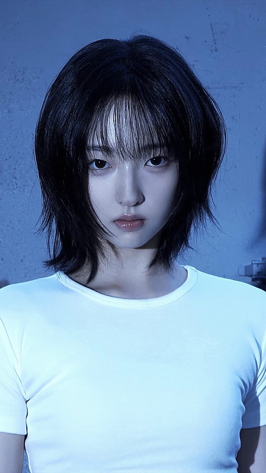 A member of A2O LITE with a layered black bob and a plain white T-shirt looks straight into the camera against a blue concrete wall in the 'NEON' music video