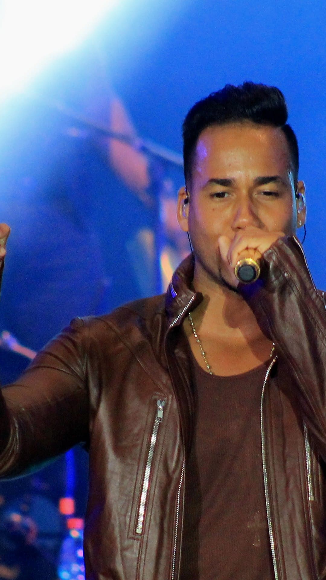 Romeo Santos, in a brown leather jacket with a silver chain, sings into a gold microphone held close to his mouth under a blue stage light at Chile's Estadio Nacional in January 2015