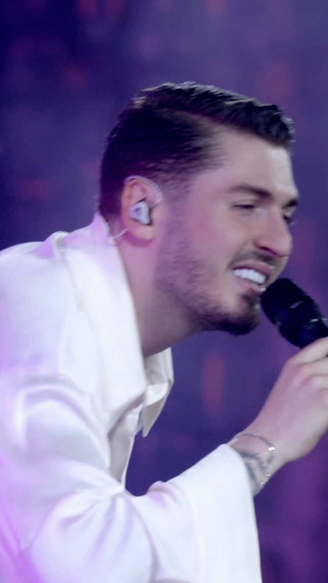 BLOK3, in a loose white shirt with an in-ear monitor, smiles as he sings 'Sebebi Yar' into a black microphone under purple stage light at Beşiktaş Tüpraş Stadium in Istanbul