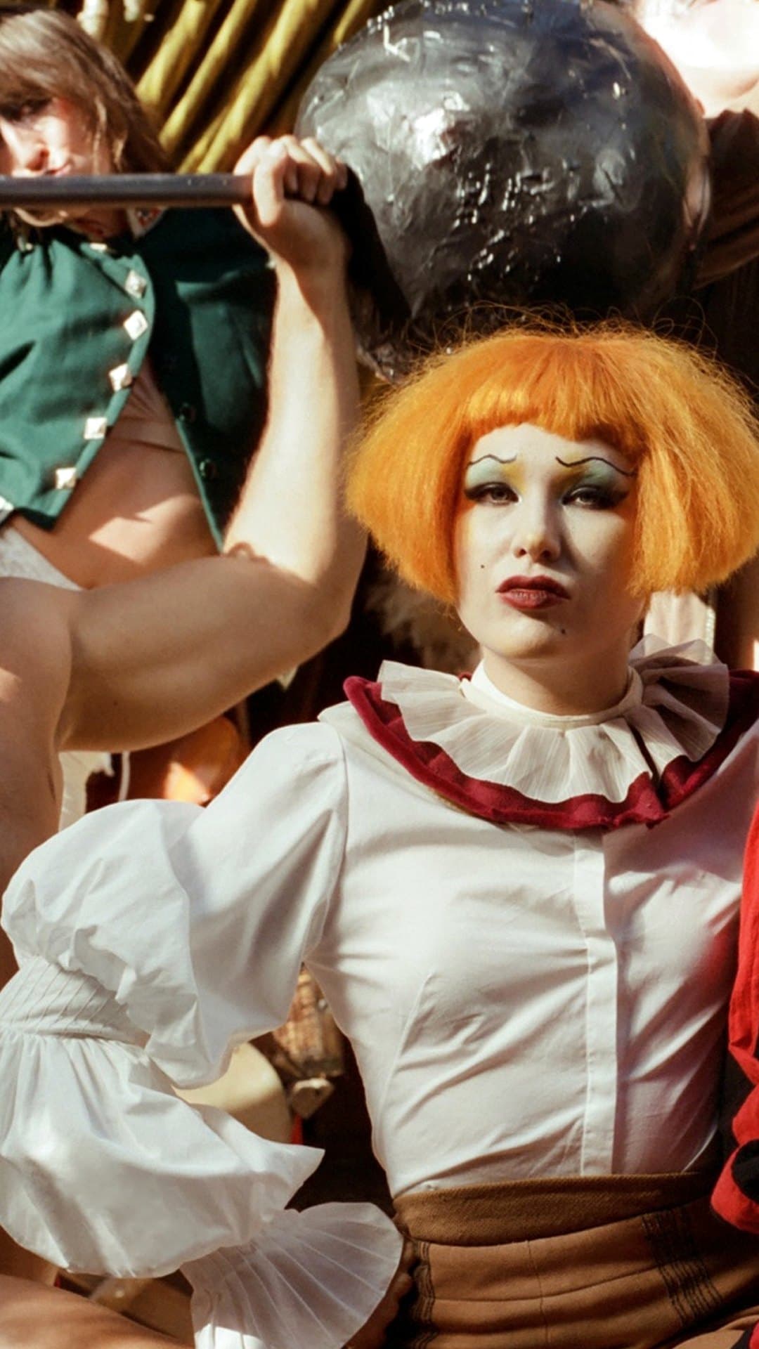 Haute, with an orange bob, white clown make-up, drawn-on brows and a ruffled white collar, looks straight at the camera in front of a circus strongman lifting a barbell, on the cover of Haute & Freddy's album 'Big Disgrace'