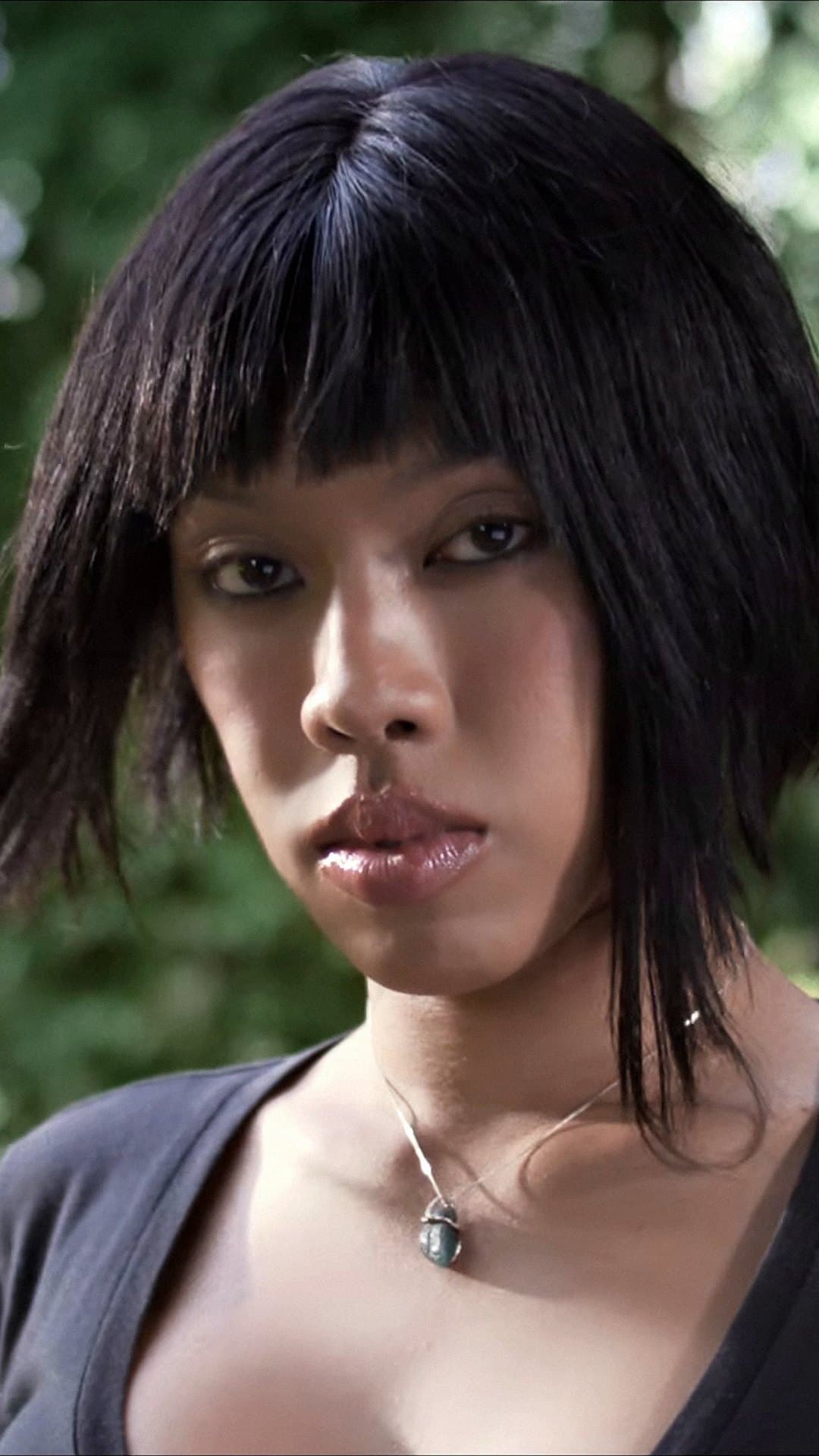 ALIE BLACKCOBRA, in a black bob with a fringe, looks straight at the camera in front of green trees in the 'SOMEONE (สรณะ)' video