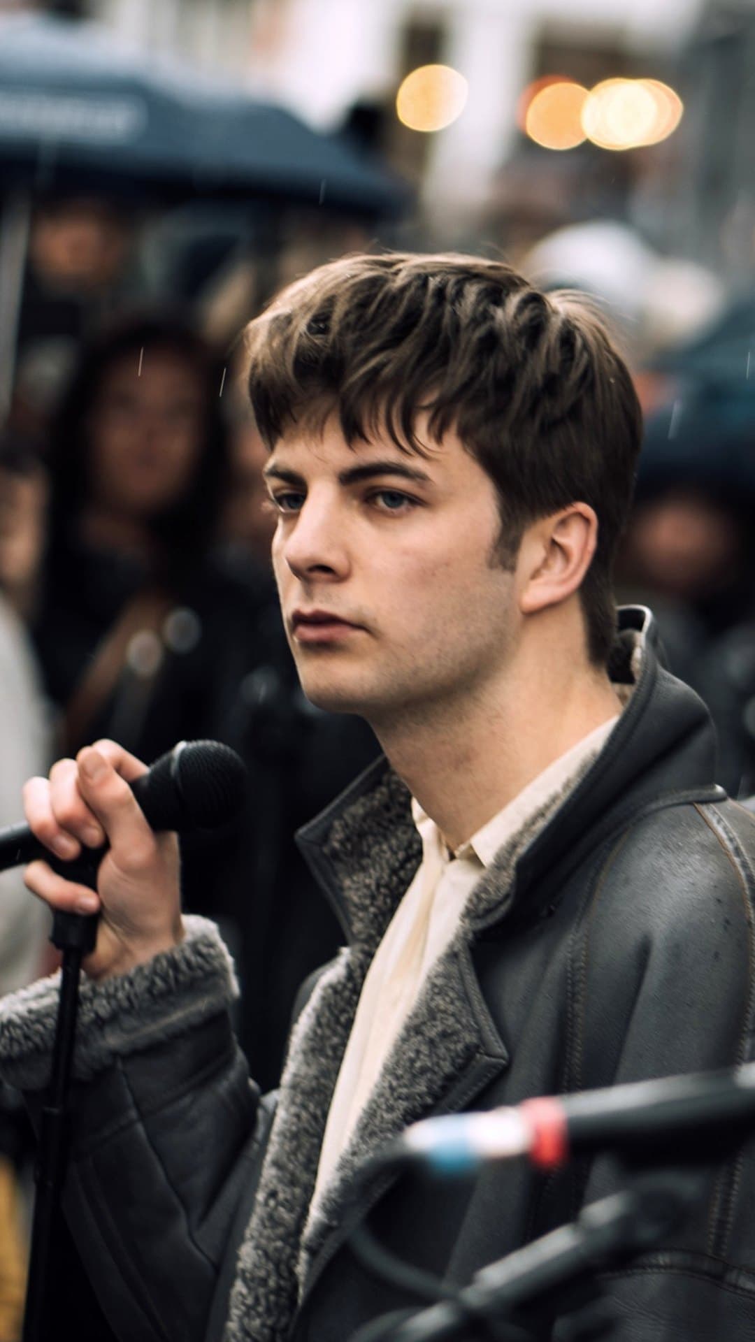 Grian Chatten of Fontaines D.C., in a grey shearling jacket, holds a microphone and looks to one side in front of a crowd under umbrellas at Rough Trade West, London, in April 2019