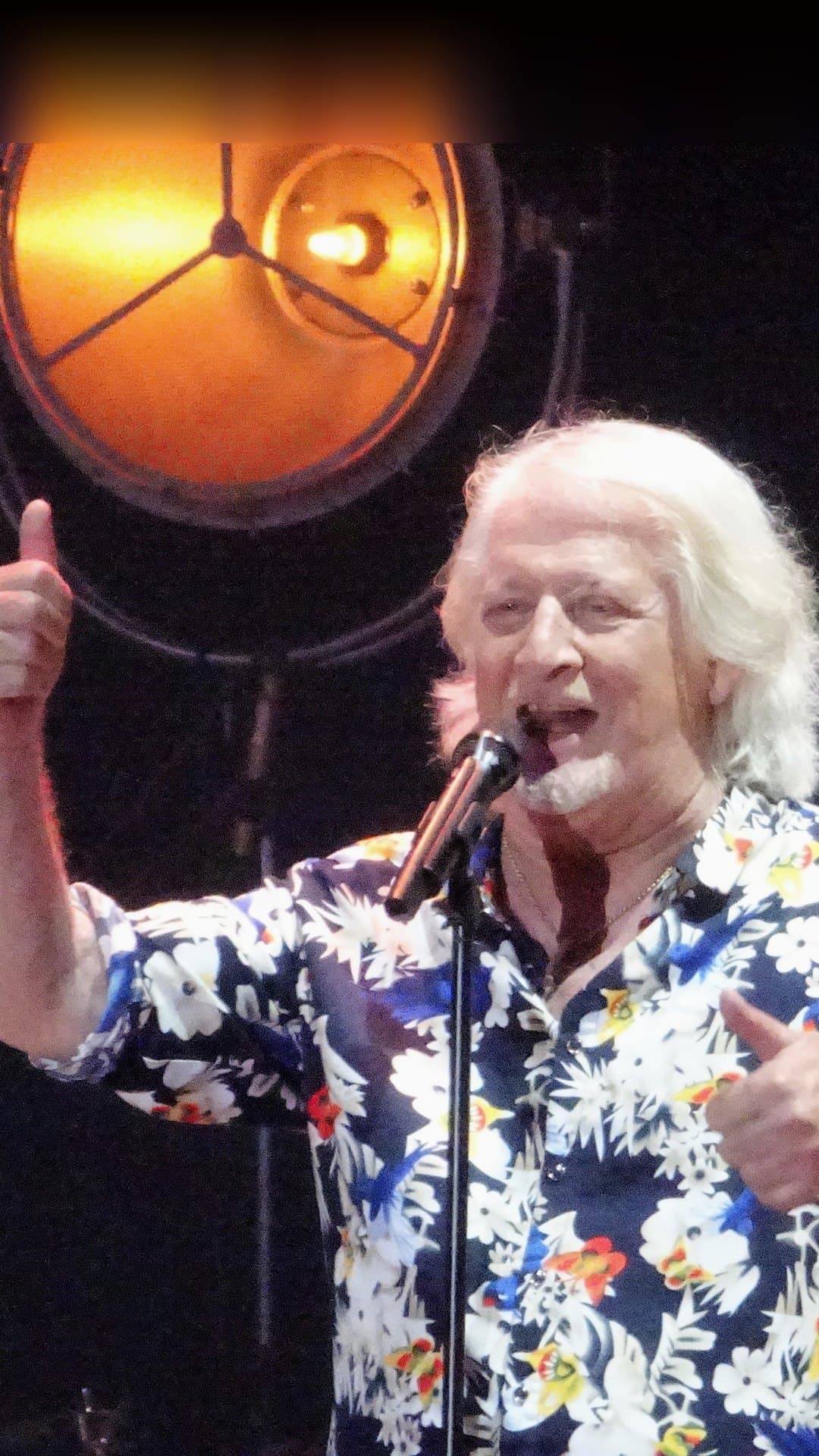 Patrick Sébastien, with long white hair and a white shirt printed with blue and yellow flowers, sings into a microphone and raises a thumb under a round orange stage lamp, in concert at Somain, France, on 2 July 2022