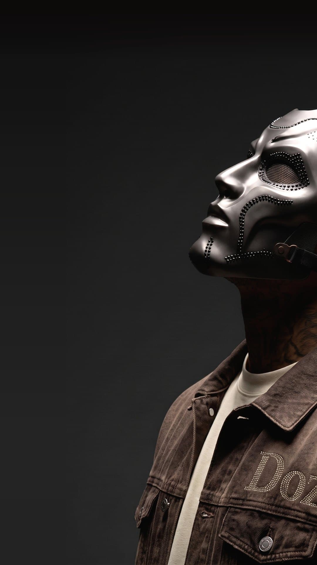 Doz in profile, looking upward in a silver metal mask studded with dark crystals, braids and a stud earring showing, against a dark backdrop, in the picture on his YouTube channel