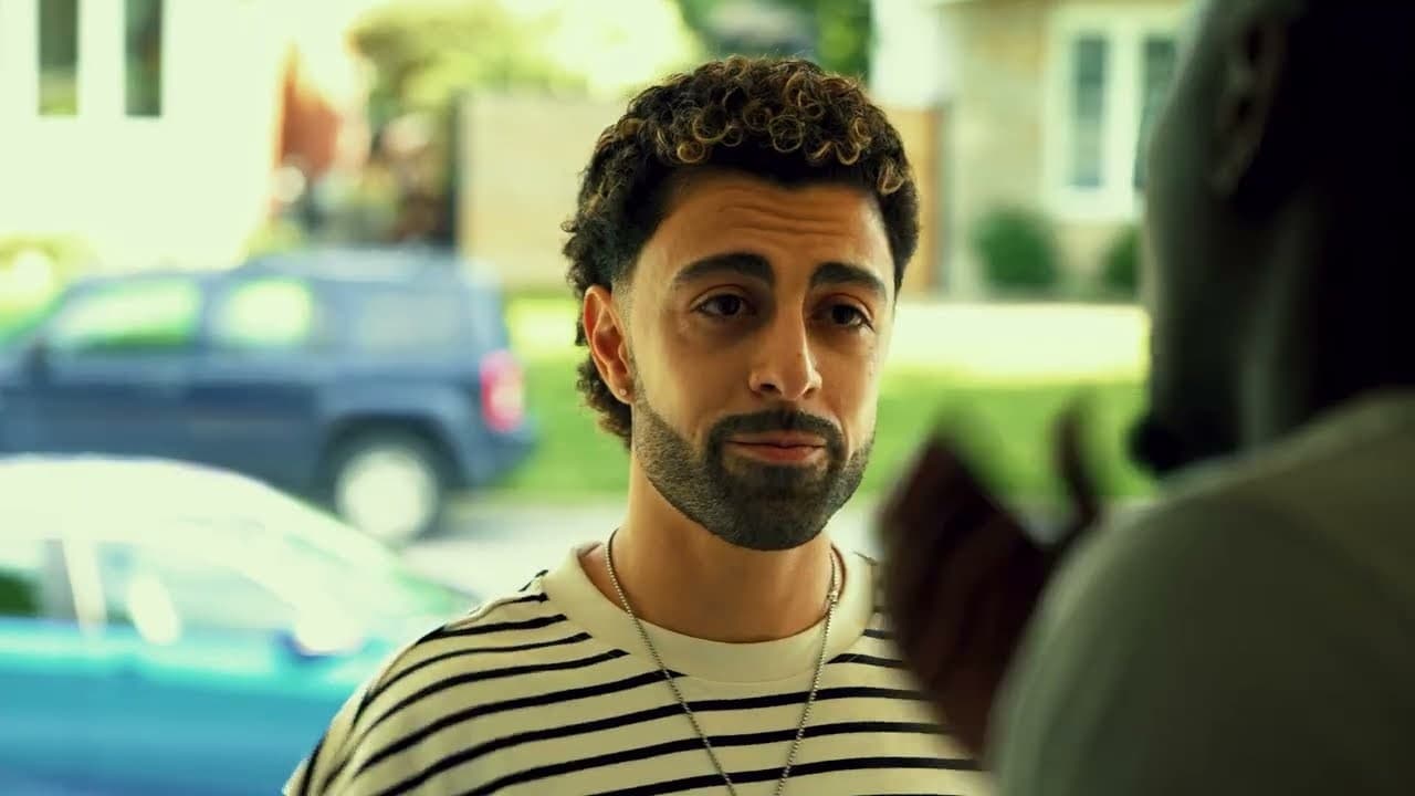 Lexnour in a striped T-shirt stands on a front path talking to a man in the foreground, cars parked behind them, in the last scene of the 'God I Need You' video