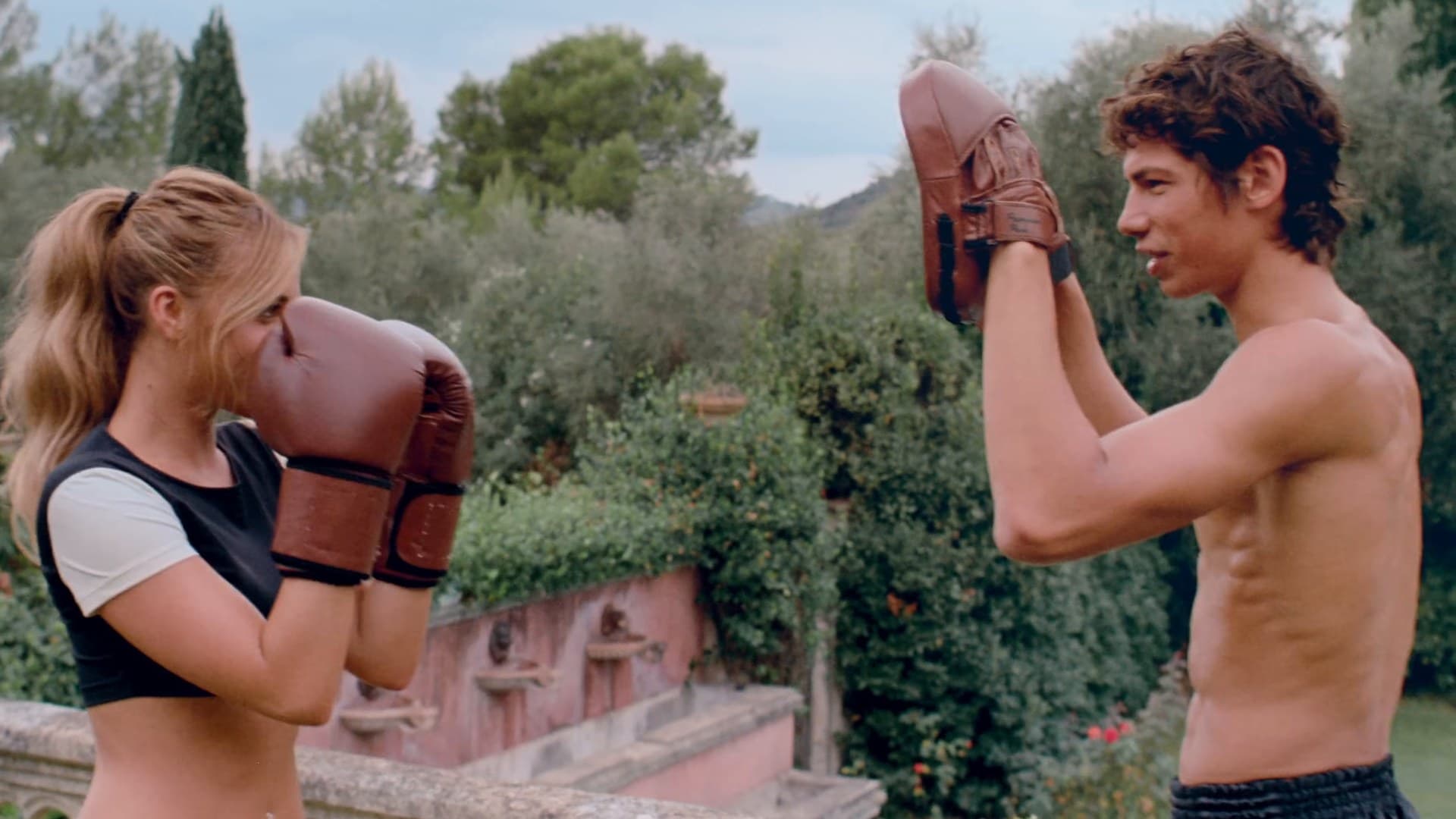 Tate McRae, in red boxing gloves and a black-and-white crop top, spars with a shirtless young man holding punch pads in a hillside garden in the 'Housewife' music video