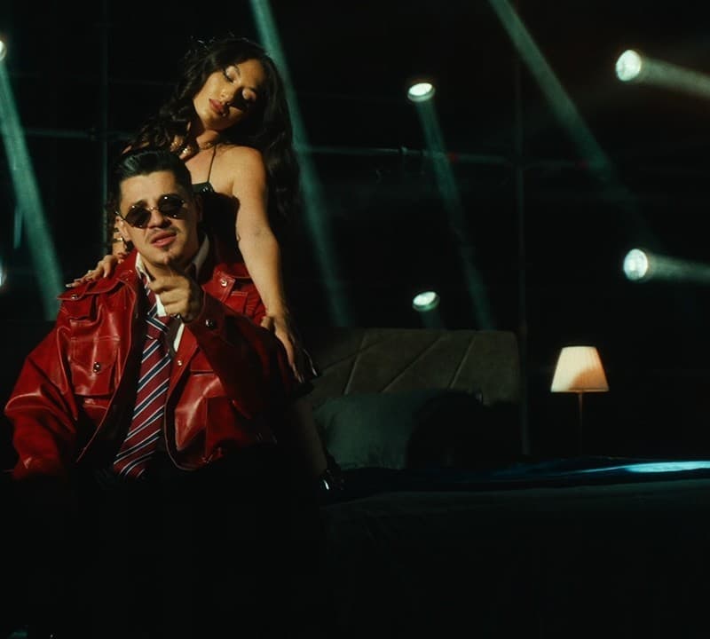 Bogdan DLP in a red leather jacket and sunglasses points at the camera while a woman leans over his shoulders, on a bedroom set lit by stage lamps, in the official video for 'În locul meu'
