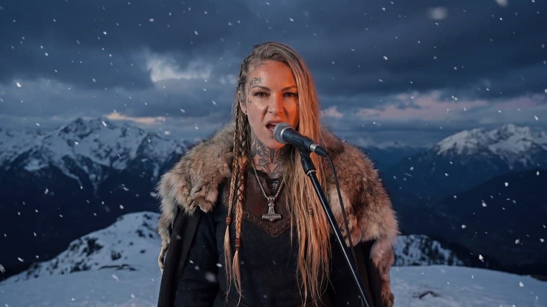 A singer in a fur cloak, with long braids and tattoos on her neck and temple, sings into a microphone on a snowy mountain top in Bjorn Bonebreaker's 'Alehouse Wolf Call' video