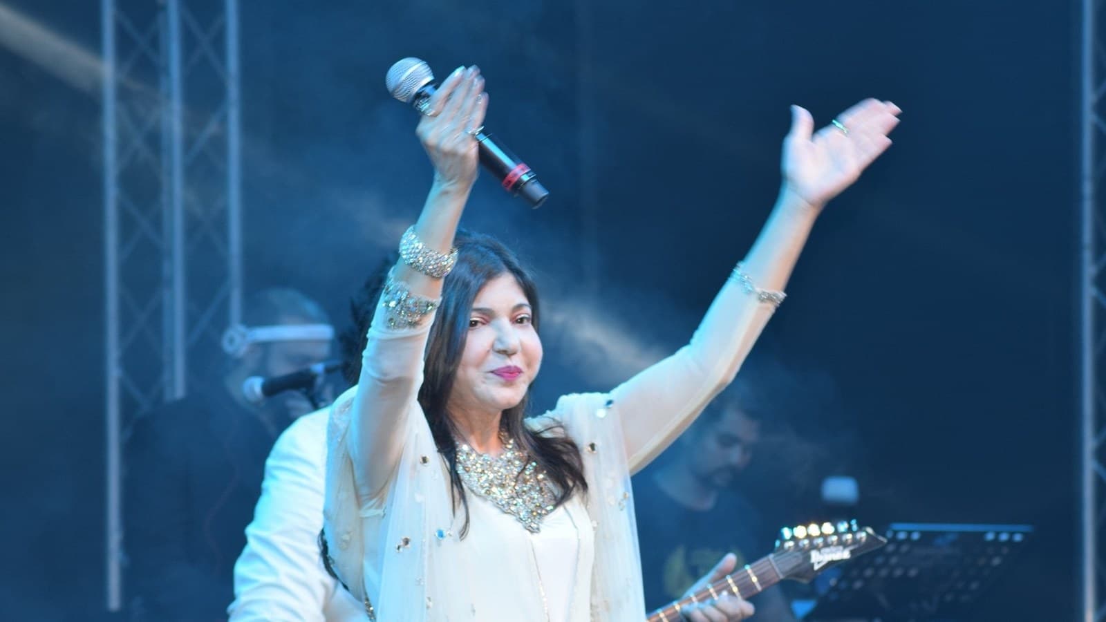 Alka Yagnik in a white outfit raises both arms, a microphone in one hand, on a concert stage in blue light, with a guitarist behind her