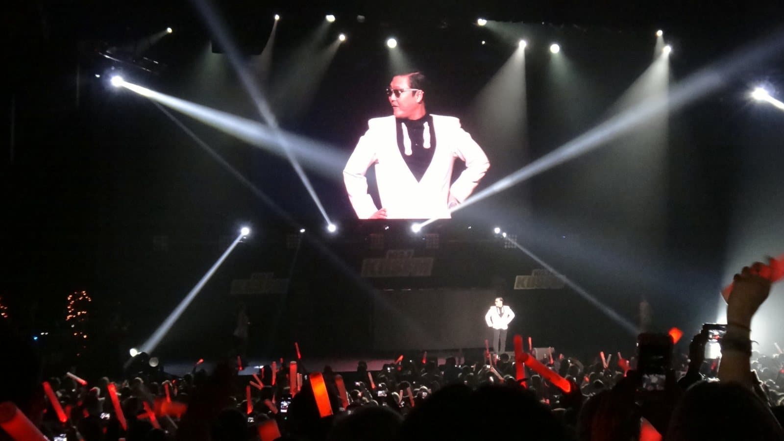 PSY, in a white jacket and dark sunglasses, appears on a giant screen above a concert stage while he performs 'Gangnam Style' below, lit by spotlights over a crowd waving red light sticks, at KIIS FM's Jingle Ball in December 2012