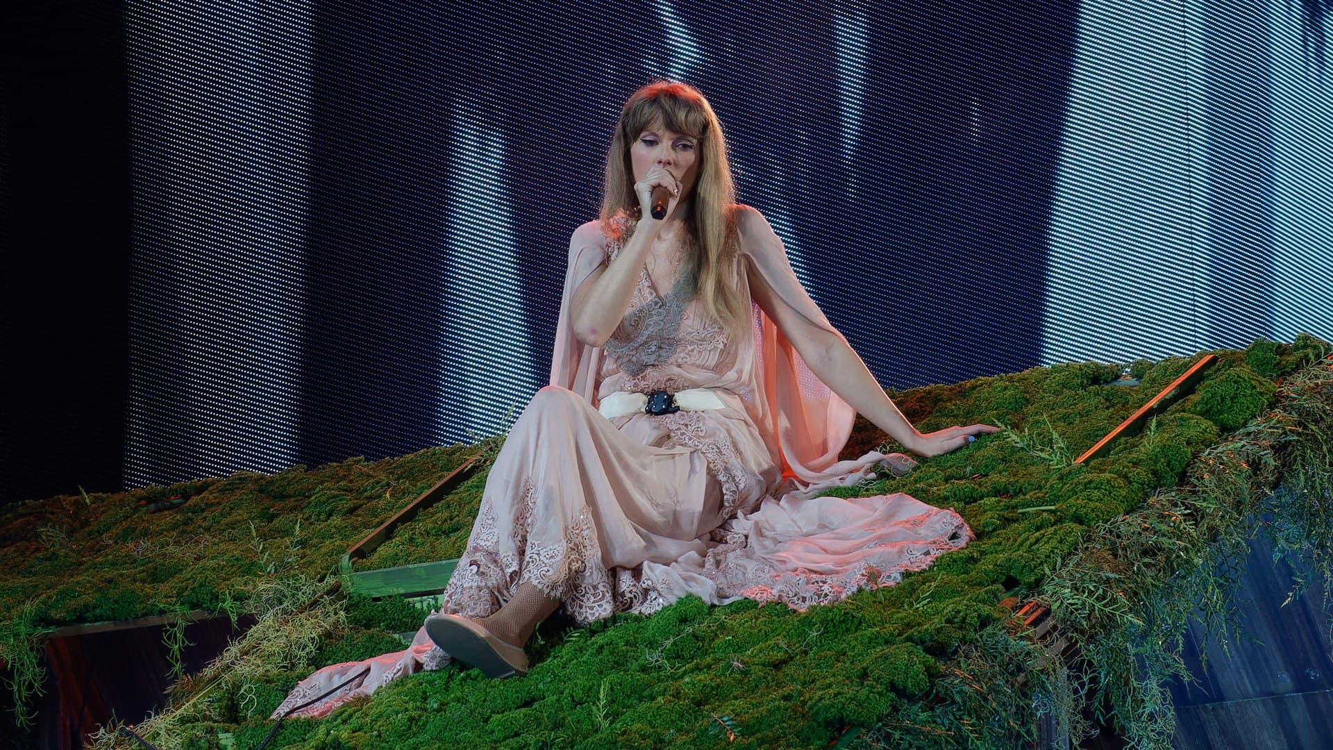 Taylor Swift in a pale pink lace gown sits on a moss-covered stage roof and sings into a microphone during the Eras Tour in Arlington, Texas, in April 2023