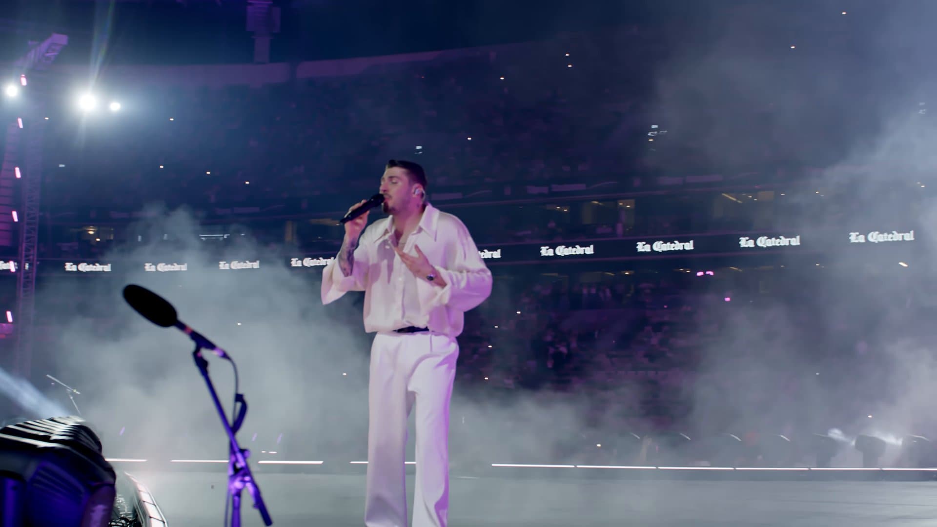BLOK3, dressed all in white, sings 'Sebebi Yar' alone on a smoky stadium stage at night, with a microphone stand in front of him and 'La Catedral' lit along the stands behind