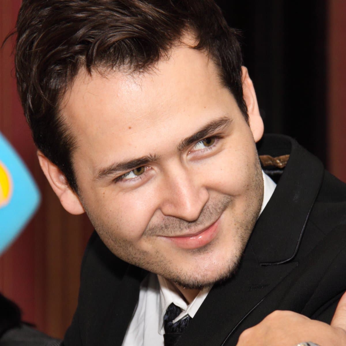 Edward Maya, dark-haired and smiling over his shoulder, in a black suit, white shirt and dark tie in front of a red curtain