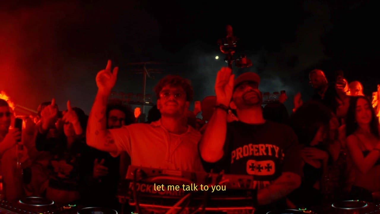 Two men behind DJ decks under red light raise their hands in front of a dancing crowd, a lyric caption at the bottom of the frame, in the official lyric video for ANOTR's 'Talk To You'
