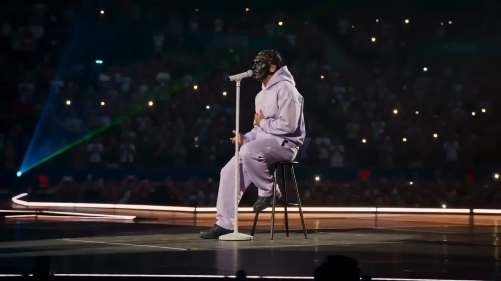 Doz, masked and in a lilac tracksuit, sits on a stool at a microphone stand on a stage before a dark stadium crowd holding up phone lights, in his video for 'BLOQUÉ'
