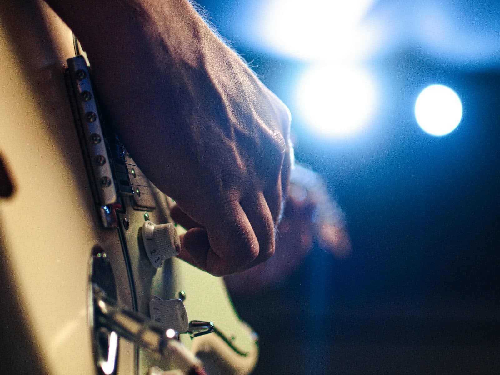 A guitarist's hand strumming a white electric guitar under blue stage lights