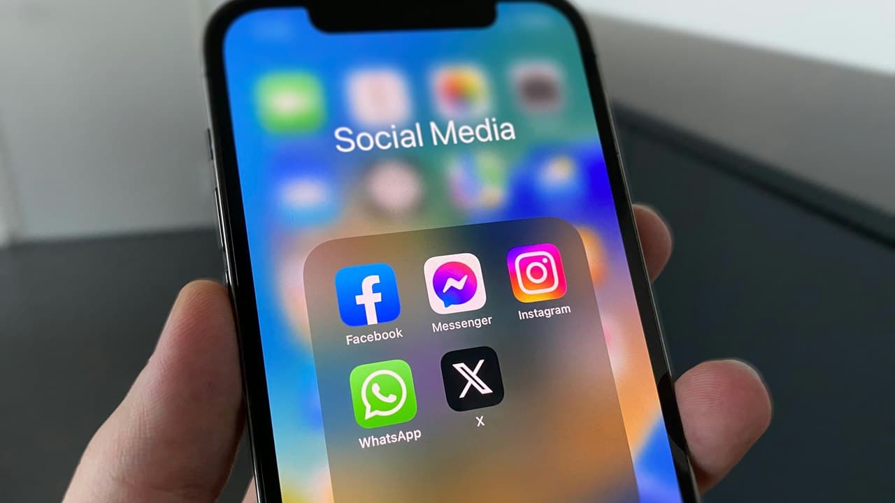 A hand holds an iPhone open to a “Social Media” folder of app icons: Facebook, Messenger, Instagram, WhatsApp and a black X icon