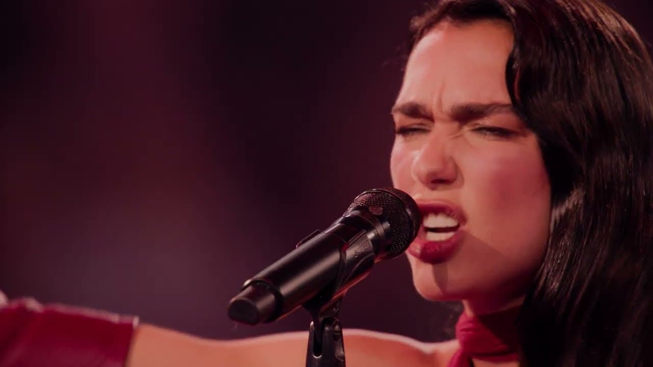 Close-up of Dua Lipa mid-chorus in the 'Training Season' live video, eyes shut, singing into a black microphone against a dark red backdrop