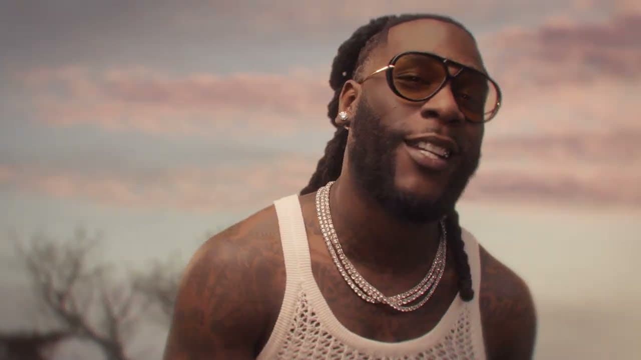Shakira & Burna Boy 'Dai Dai' MV capture: Burna Boy in sunglasses and a white mesh vest with a silver chain smiles at dusk