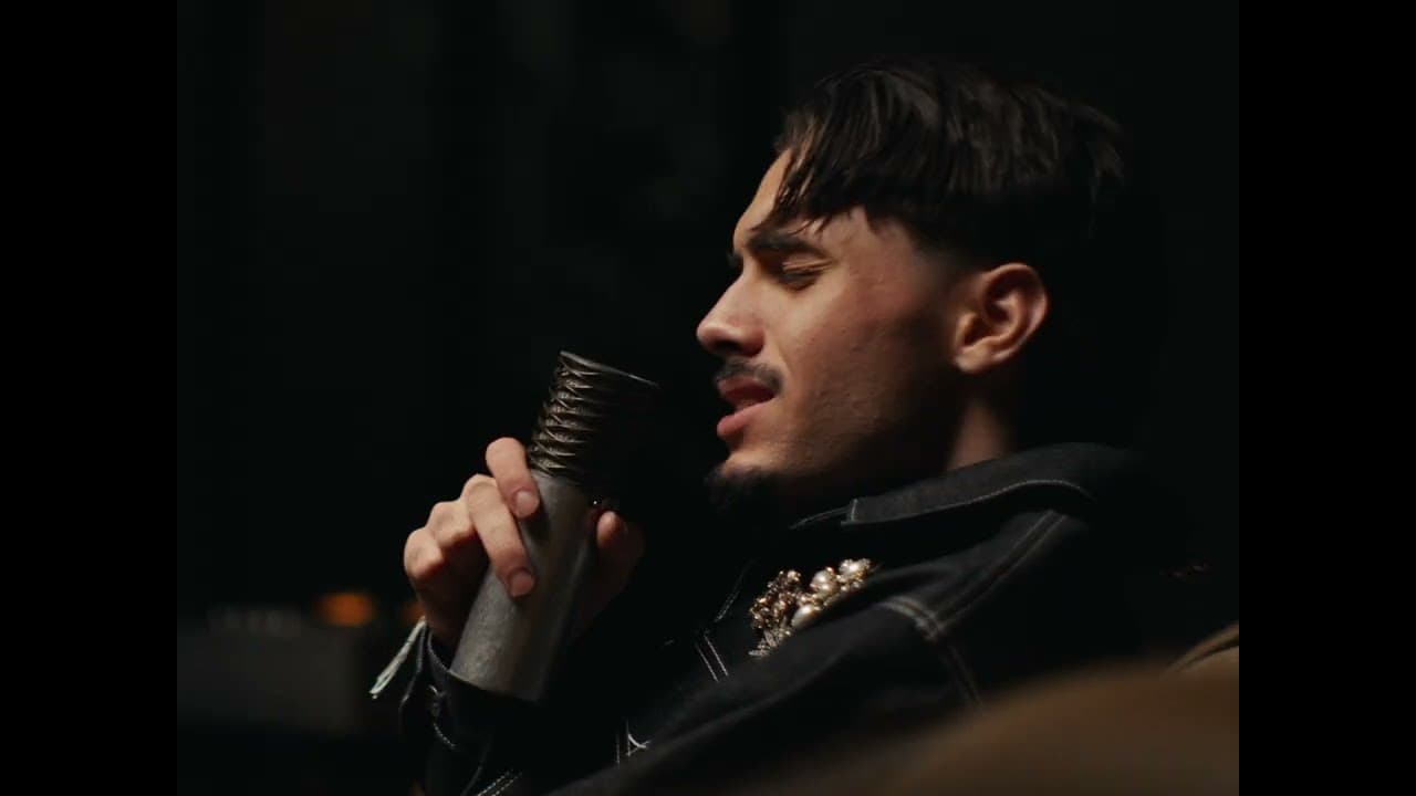 RnBoi, eyes closed, sings into an old-style silver microphone in a dark room, wearing a denim jacket with a jewelled brooch, in the 'J'assume' clip